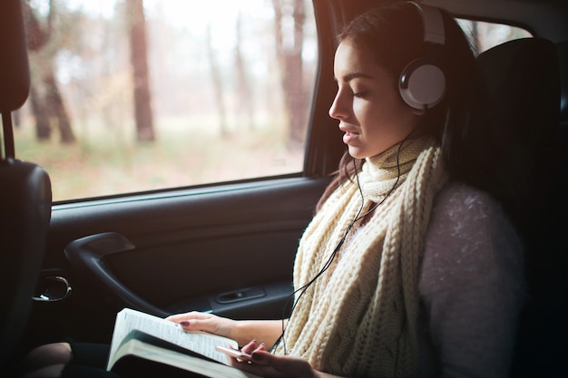 Femme dans la voiture, concept automne. Jolie fille souriante, écouter de la musique avec des écouteurs et lire un livre se déplaçant en voiture.