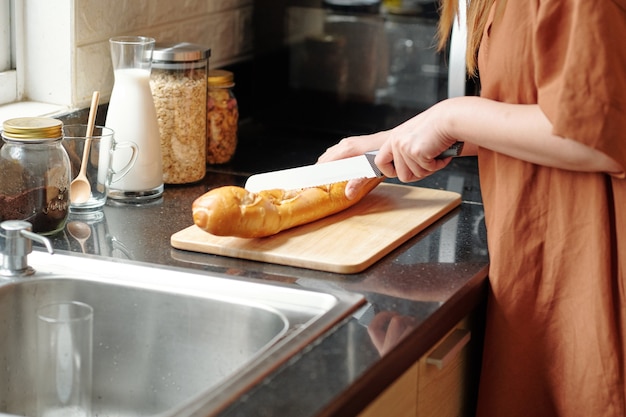 Femme debout au comptoir de la cuisine et coupant une baguette fraîche avec un couteau tranchant
