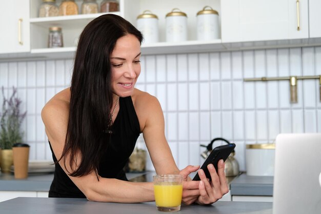 Une femme debout dans la cuisine boit du jus dans un verre et communique en ligne par téléphone