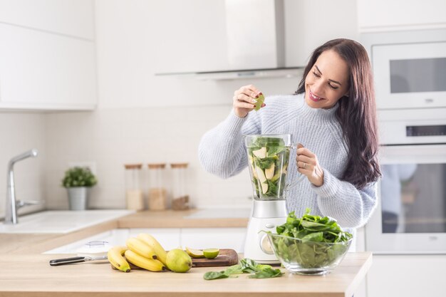 Femme debout dans une cuisine pressant du citron vert dans un mélangeur plein de fruits et légumes.