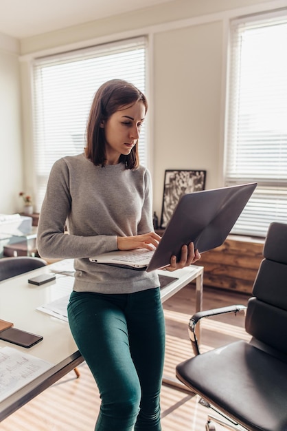 Femme debout près d'un bureau avec un ordinateur portable à la main