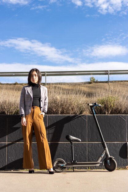 Femme décontractée intelligente à côté d'un scooter électrique sur le trottoir Jeune femme près d'un scooter électrique regardant la caméra