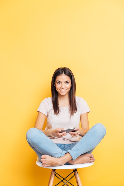 Femme décontractée souriante assise sur la table et utilisant un smartphone sur un mur jaune