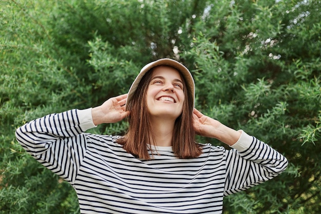 Femme décontractée souriante portant une chemise rayée et un panama debout avec les bras levés près d'un buisson vert dans le parc profitant de l'été et marchant dehors par beau temps