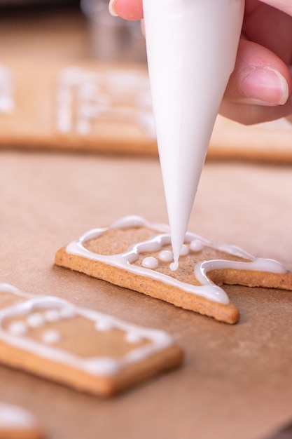 Photo une femme décore une maison de biscuits en pain d'épice avec une garniture de glaçage blanc sur fond de table en bois, du papier sulfurisé dans la cuisine, gros plan, macro.