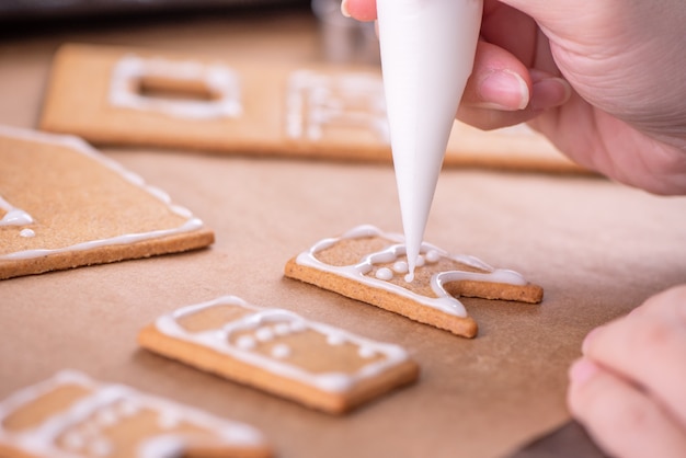 Une femme décore une maison de biscuits en pain d'épice avec une garniture de glaçage blanc sur fond de table en bois, du papier sulfurisé dans la cuisine, gros plan, macro.