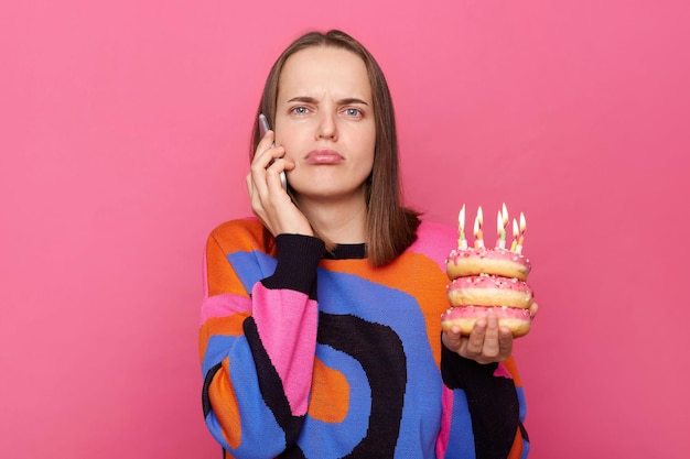 Femme déçue tenant un gâteau savoureux avec des bougies allumées porte un pull décontracté célébrant l'anniversaire seul en attendant que les invités parlent au téléphone avec une expression triste isolée sur fond de studio gris