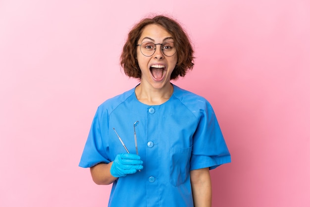 Femme dentiste anglais tenant des outils sur isolé sur un mur rose avec une expression faciale surprise