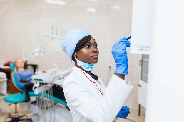 Une femme dentiste noire dans un cabinet dentaire parlant avec une patiente et se préparant au traitement étudie une radiographie. Équipement médical moderne