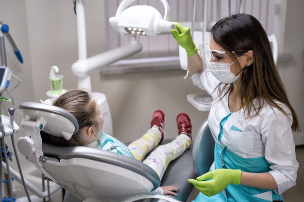 Femme dentiste préparant la lumière pour le traitement à la clinique dentaire. Elle a une petite fille patiente.