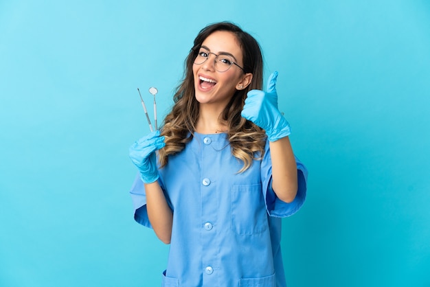 Femme dentiste tenant des outils sur isolé sur mur bleu avec les pouces vers le haut parce que quelque chose de bien s'est passé