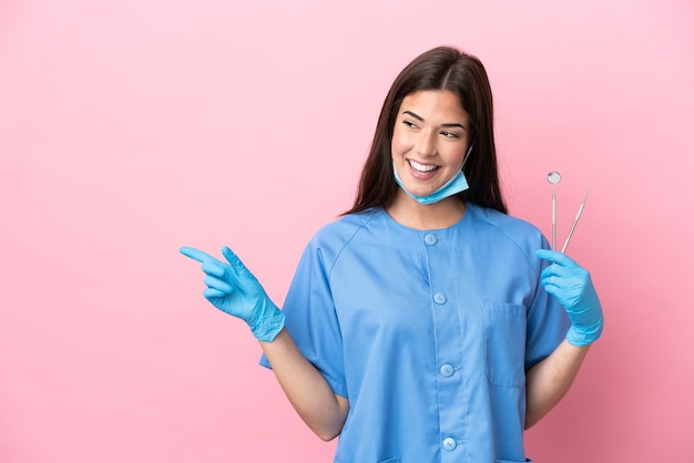 Femme dentiste tenant des outils isolés sur fond rose, pointant le doigt sur le côté et présentant un produit