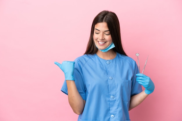 Femme dentiste tenant des outils isolés sur fond rose pointant vers le côté pour présenter un produit