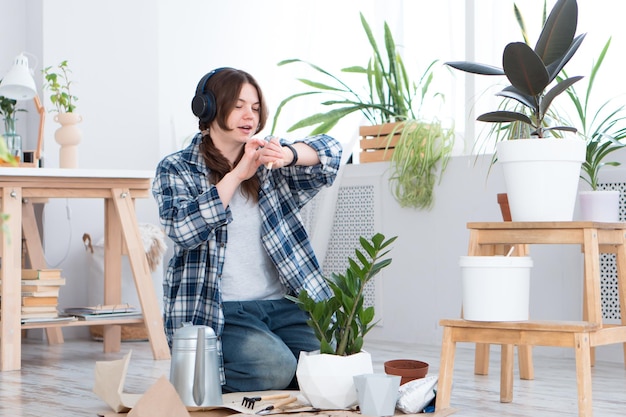 Femme déplaçant une plante dans un nouveau pot et écoutant de la musique à l'aide d'écouteurs sans fil et d'une montre intelligente à la maison