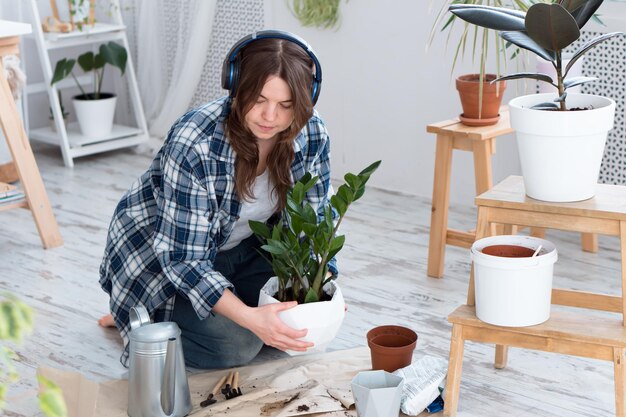 Une femme déplace une plante de zamiokulkas dans un nouveau pot et écoute de la musique à l'aide d'un casque sans fil à la maison