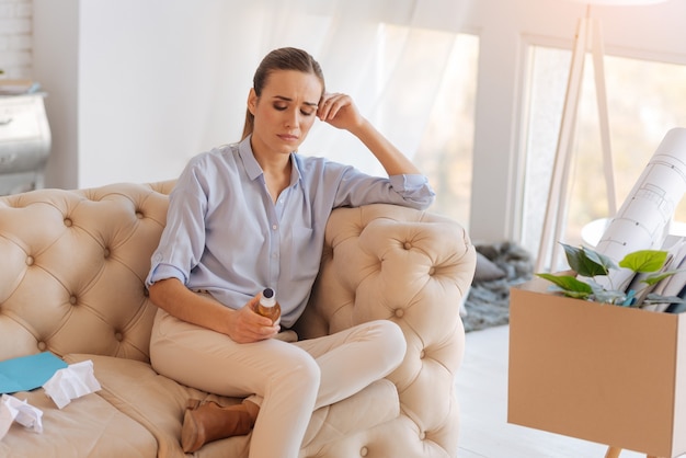 Femme déprimée. Femme au chômage nerveuse émotionnelle surmontant sa dépression avec une aide d'alcool tandis qu'une boîte avec de vieux schémas et des graphiques debout non loin d'elle