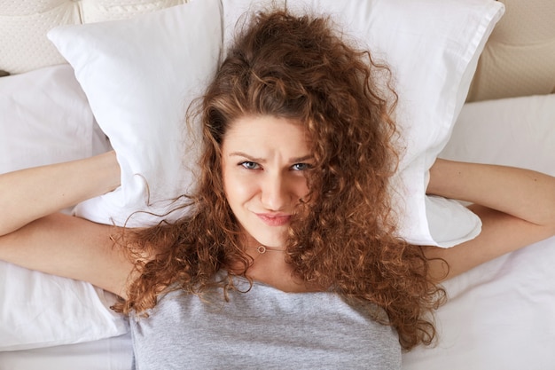 Une femme déprimée frustrée couvre les oreilles avec un oreiller pendant qu'elle se trouve au lit, ne supporte pas le ronflement de quelqu'un, ne peut pas s'endormir