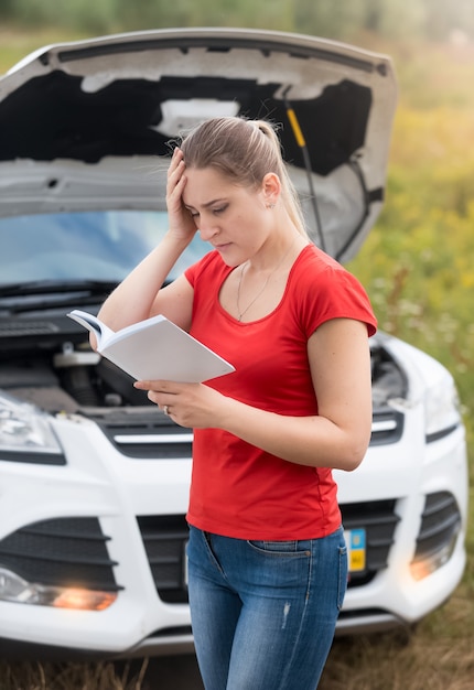 Femme déprimée regardant sa voiture cassée et lisant le manuel du propriétaire de la voiture