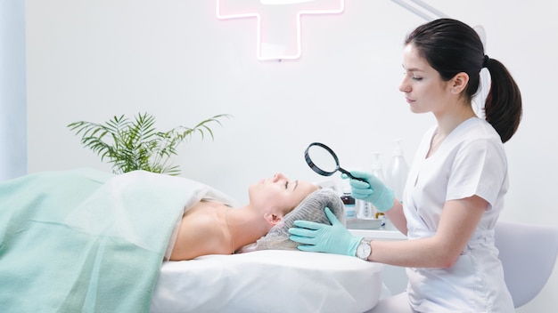 La femme dermatologue examinant le visage du jeune patient avec une loupe dans une clinique ou un salon de beauté. Concept de nettoyant pour le visage, la peau et les soins de santé