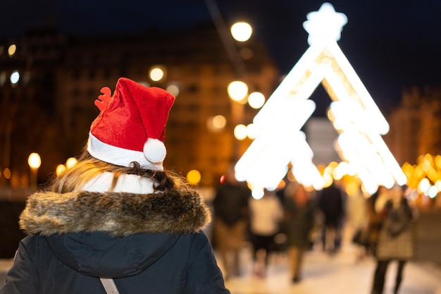 Femme de derrière avec un chapeau de noël dans la ville la nuit en hiver marchant en regardant l'éclairage