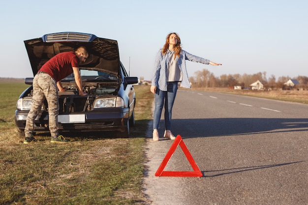 Une femme désespérée arrête les véhicules sur la route selon les besoins, voyage avec son mari en voiture et a des dommages avec le moteur