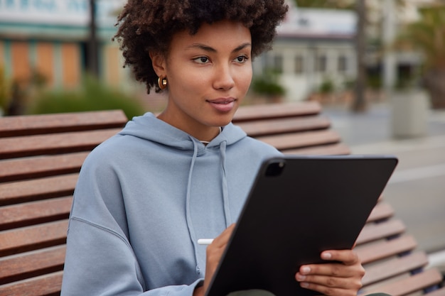 une femme dessine des croquis sur une tablette avec un stylet fait des travaux d'image sur un futur projet vêtue de poses de sweat-shirt sur un banc en bois sur la ville utilise un appareil moderne