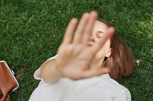 Femme détendue portant un t-shirt blanc allongé sur l'herbe verte dans le parc, tendant la main couvrant son visage avec la paume, profitant de la tome gratuite et du temps chaud de l'été