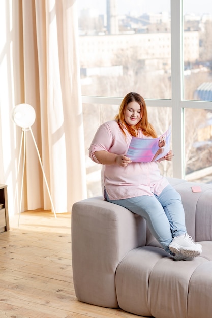 Femme dodue positive lisant sur les tendances de la mode moderne assise sur le canapé