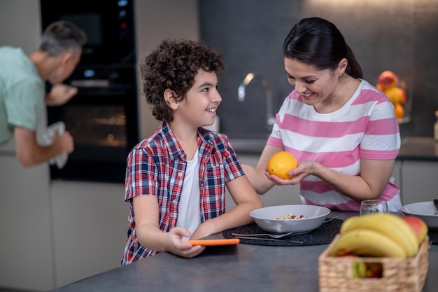 Femme donnant une orange à un garçon souriant dans la cuisine