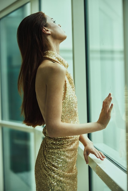 Femme dramatique dans une robe de soirée scintillante de créateur debout près de la fenêtre donnant sur la robe de soirée de l'hôtel de ville