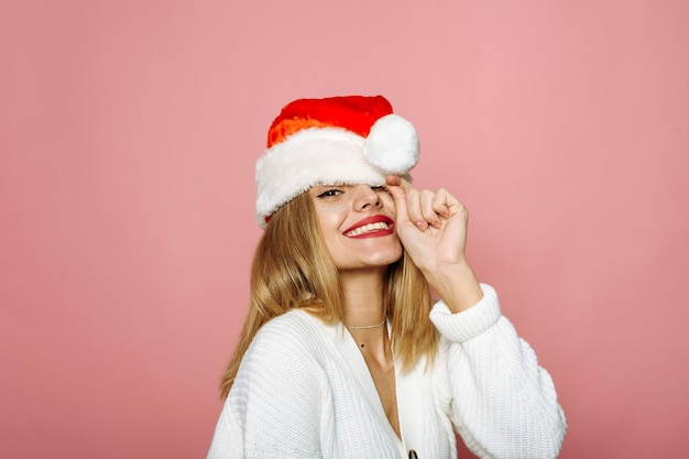 Une femme drôle couvre un œil avec un bonnet de Noel et rit. La dame coquette sur fond rose célèbre Noël et le nouvel an