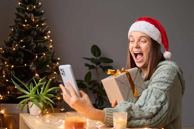 Femme drôle et joyeuse portant une chemise tricotée et un chapeau de Père Noël tenant une boîte à cadeaux assise à la maison avec une décoration de Noël célébrant la fête en ligne par appel vidéo criant d'humeur festive