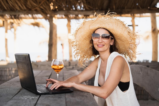 Une femme du millénaire est assise à une table dans un café tropical avec des lunettes de soleil et un chapeau de paille travaille sur un ordinateur portable et regarde la caméra