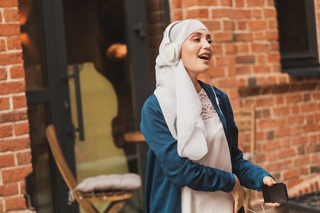 Femme du Moyen-Orient en hijab écoutant de la musique avec des écouteurs et dansant à l'extérieur. Concept d'indépendance de la femme et de féminisme.