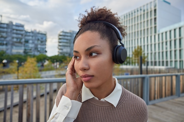 la femme écoute les paroles des chansons dans les écouteurs aime la liste de lecture préférée regarde pensivement en avant porte des vêtements décontractés pose contre la ville floue