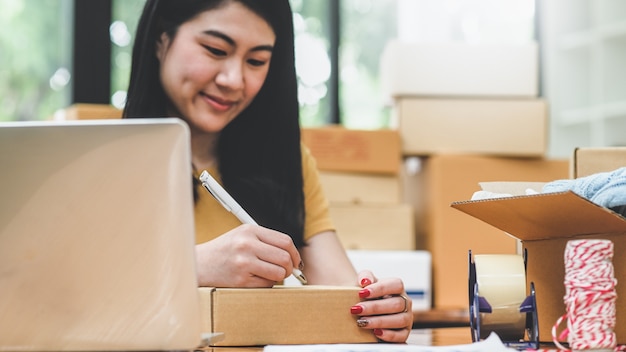 Une femme écrit une adresse sur une boîte à colis pour la livraison à un client, vend des produits en ligne, transporte des colis.