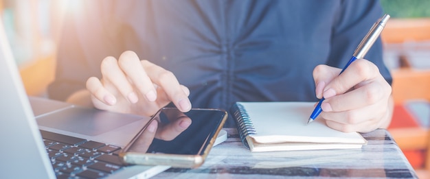 Femme écrit sur un cahier avec un stylo et elle utilise un téléphone portable au bureau. Bannière Web.