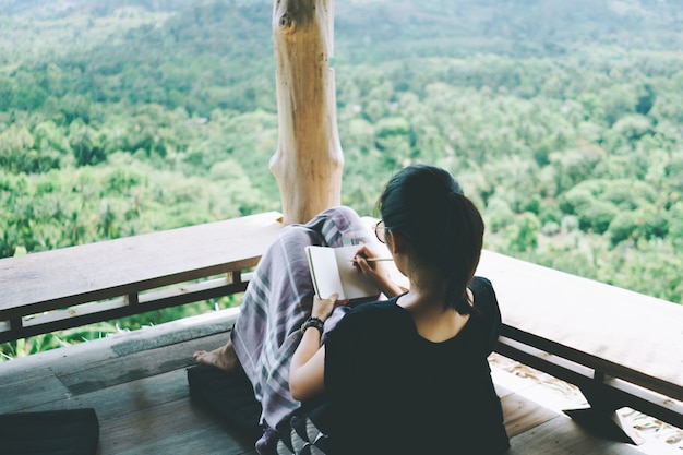 La femme écrit dans un petit carnet blanc pour prendre une note à ne pas oublier, prévoyez de faire ou d'écrire un livre avec une belle terrasse de maison et une vue sur la montagne de la nature.