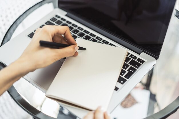 Une femme écrit avec un stylo dans un bloc-notes sur un clavier d'ordinateur portable dans un bureau ensoleillé concept d'affaires et d'éducation Close Up