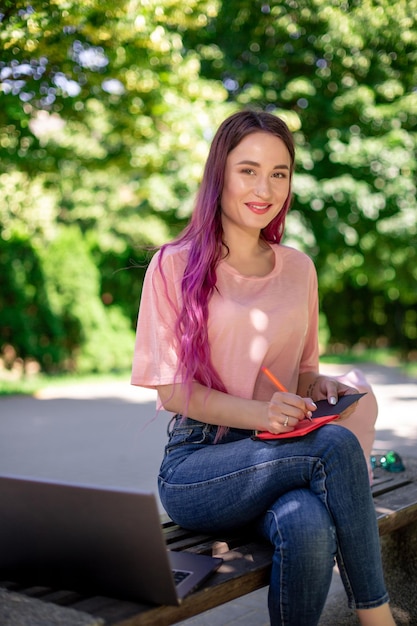 Femme écrivant dans un cahier assis sur un banc en bois dans le parc. Fille travaillant à l'extérieur sur un ordinateur portable, copiez l'espace.