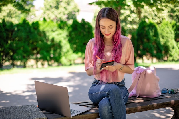 Femme écrivant Dans Un Cahier Assis Sur Un Banc En Bois Dans Le Parc. Fille Travaillant à L'extérieur Sur Un Ordinateur Portable, Copiez L'espace.