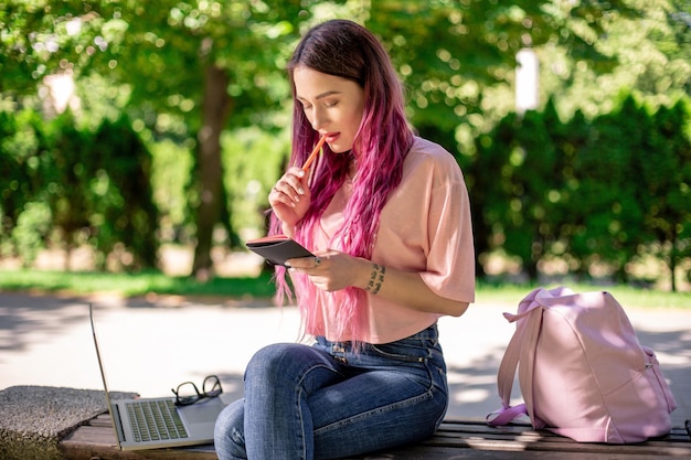 Femme écrivant dans un cahier assis sur un banc en bois dans le parc. Fille travaillant à l'extérieur sur un ordinateur portable, copiez l'espace.