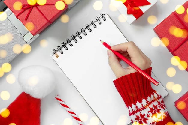 Femme écrivant dans un cahier vide sur un bureau blanc Mains de fille avec un crayon parmi une boîte-cadeau rouge et un décor festif d'hiver Concept de journal intime Salutations de vacances ou planification Vue de dessus Mock up Paper Blank