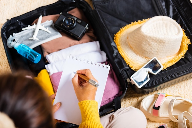 Photo femme écrivant du papier prendre note et emballer la valise pour la liste de papier à lettres de vacances assis sur la chambre