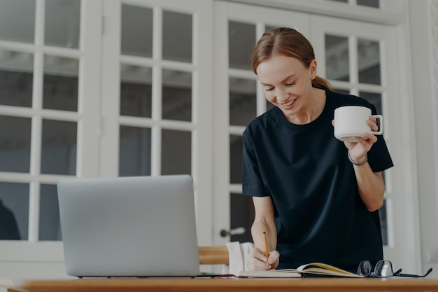 Femme écrivant des notes dans un cahier debout au bureau tenant une tasse de café planifiant le calendrier sur le lieu de travail