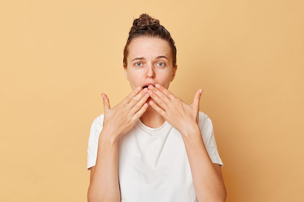 Photo une femme effrayée et choquée portant un t-shirt blanc lave les cheveux debout avec de la mousse de shampoing sur la tête isolée sur fond beige à l'aide d'un masque pour hydrater les cheveux