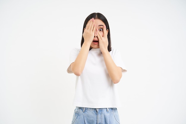 Une femme effrayée ferme les yeux, regarde à travers les doigts avec une expression de visage effrayée et choquée, dos blanc