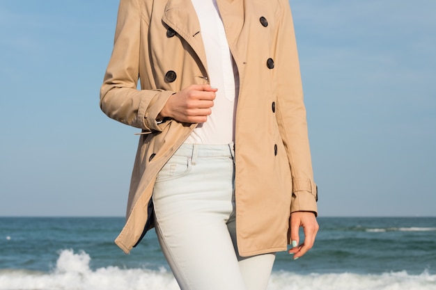 Femme élancée en manteau et jeans marchant le long de la côte sous le soleil du soir