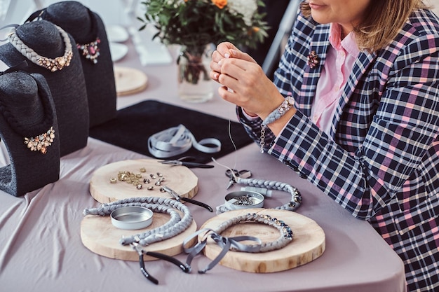 Une femme élégamment vêtue fabrique des colliers faits à la main, enfilant une aiguille dans un atelier de bijoux.