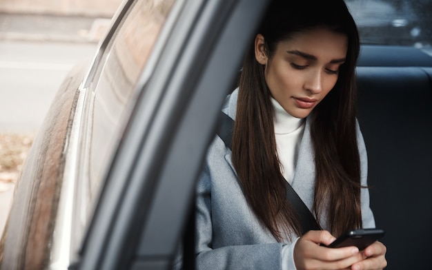 Femme élégante assise sur la banquette arrière de sa voiture et lire un message sur téléphone mobile